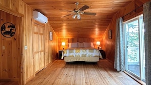 Een slaapkamer in een houten blokhut met een bed, twee bedlampjes, een plafondventilator en een raam met gordijnen.