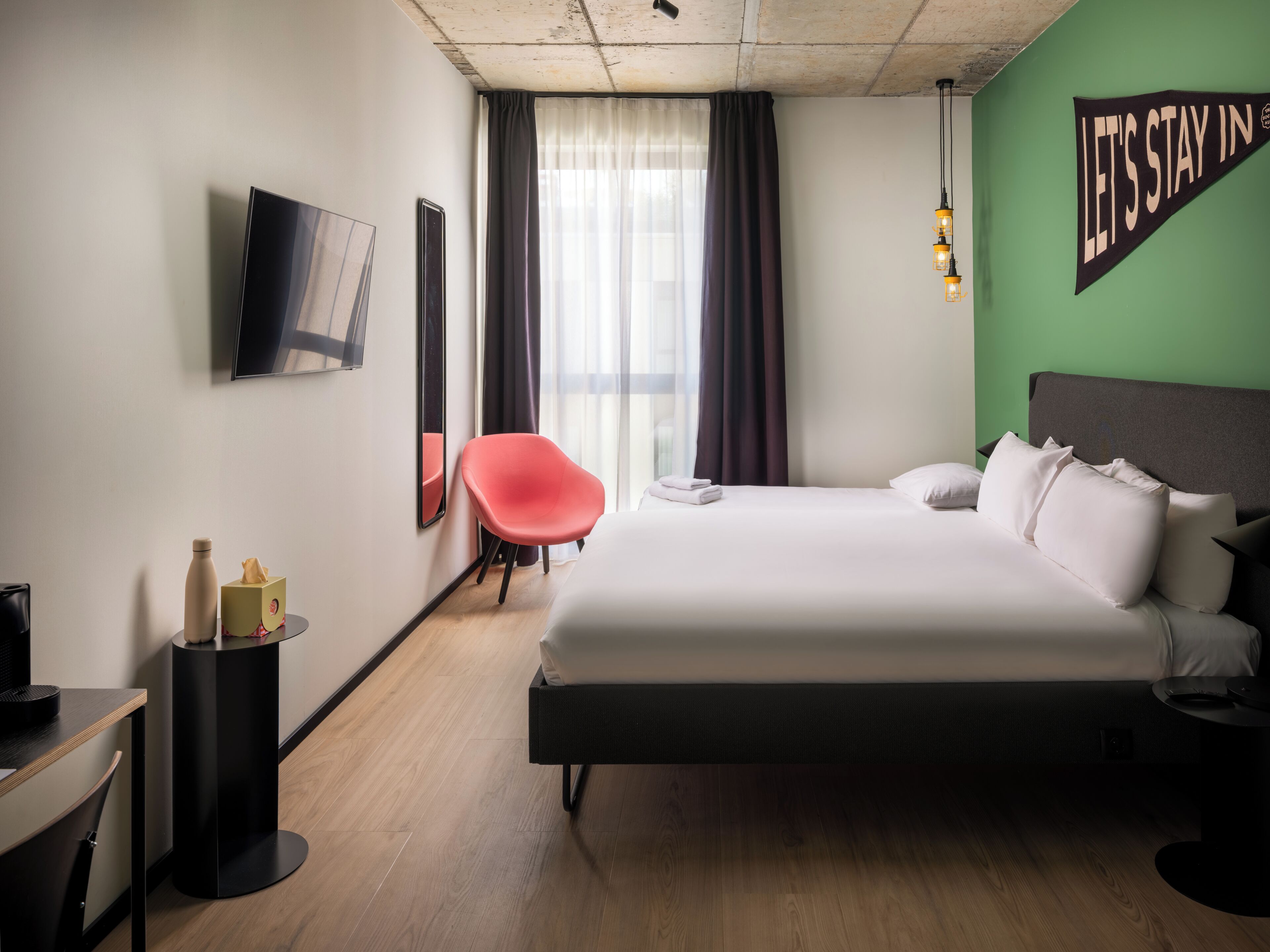 A hotel room with a bed, a chair, a TV, and a wall with a message.
