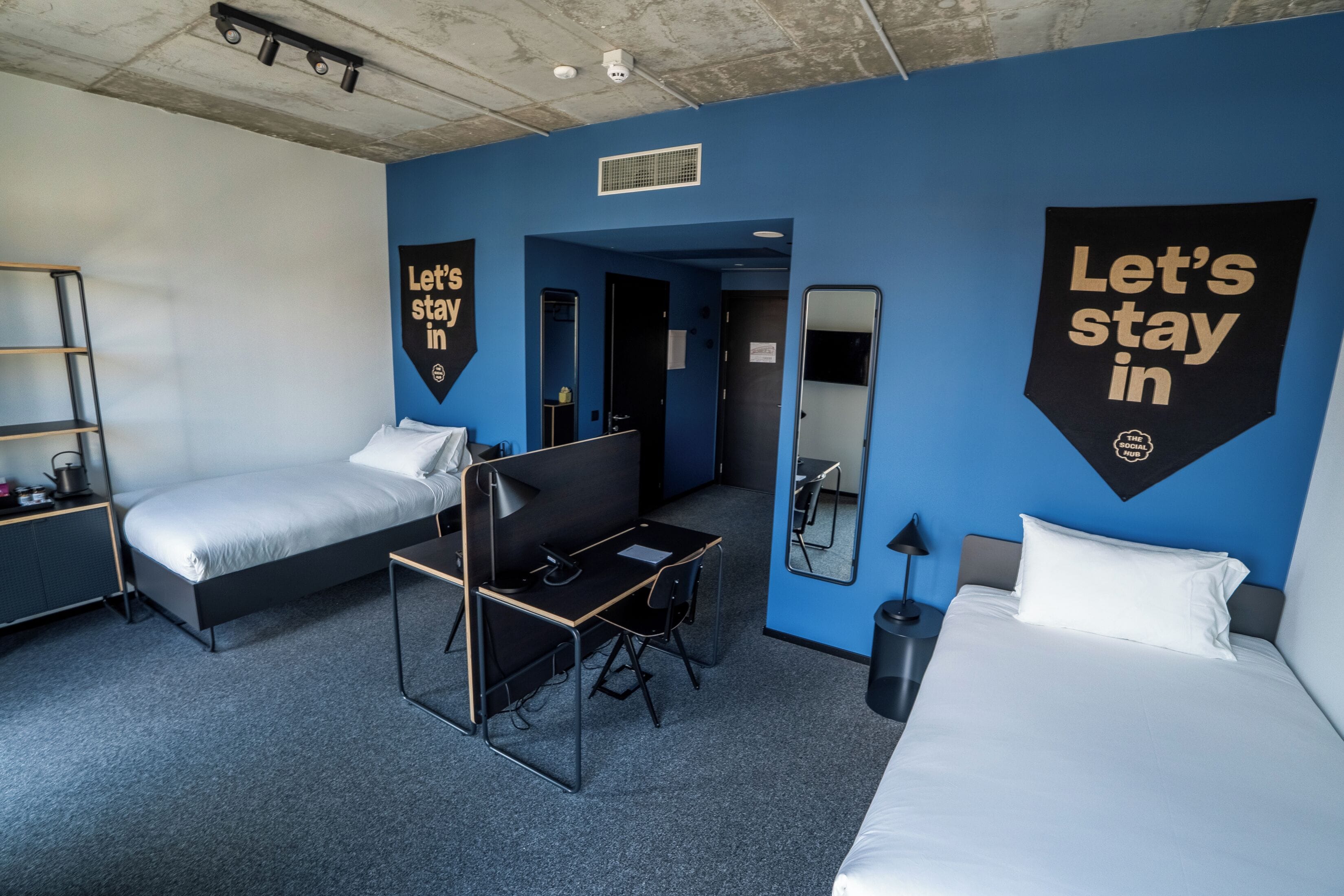 A hotel room with a bed, a desk, a chair, a wardrobe, and a mirror. There are two large banners on the walls with the text "Let's stay in".