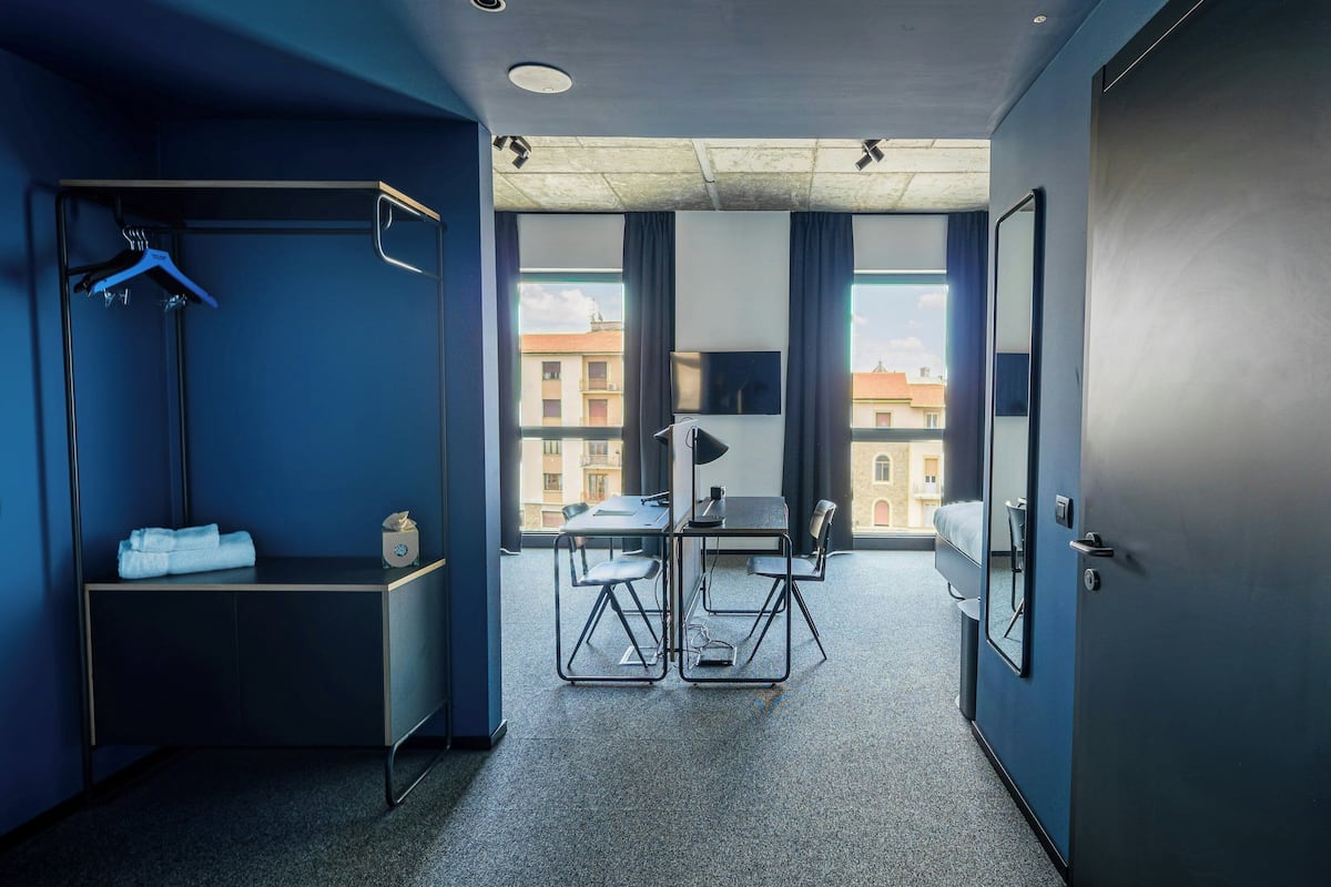 A hotel room with a bed, a desk, a chair, a wardrobe, a hanging rack, and a window with a view of buildings.