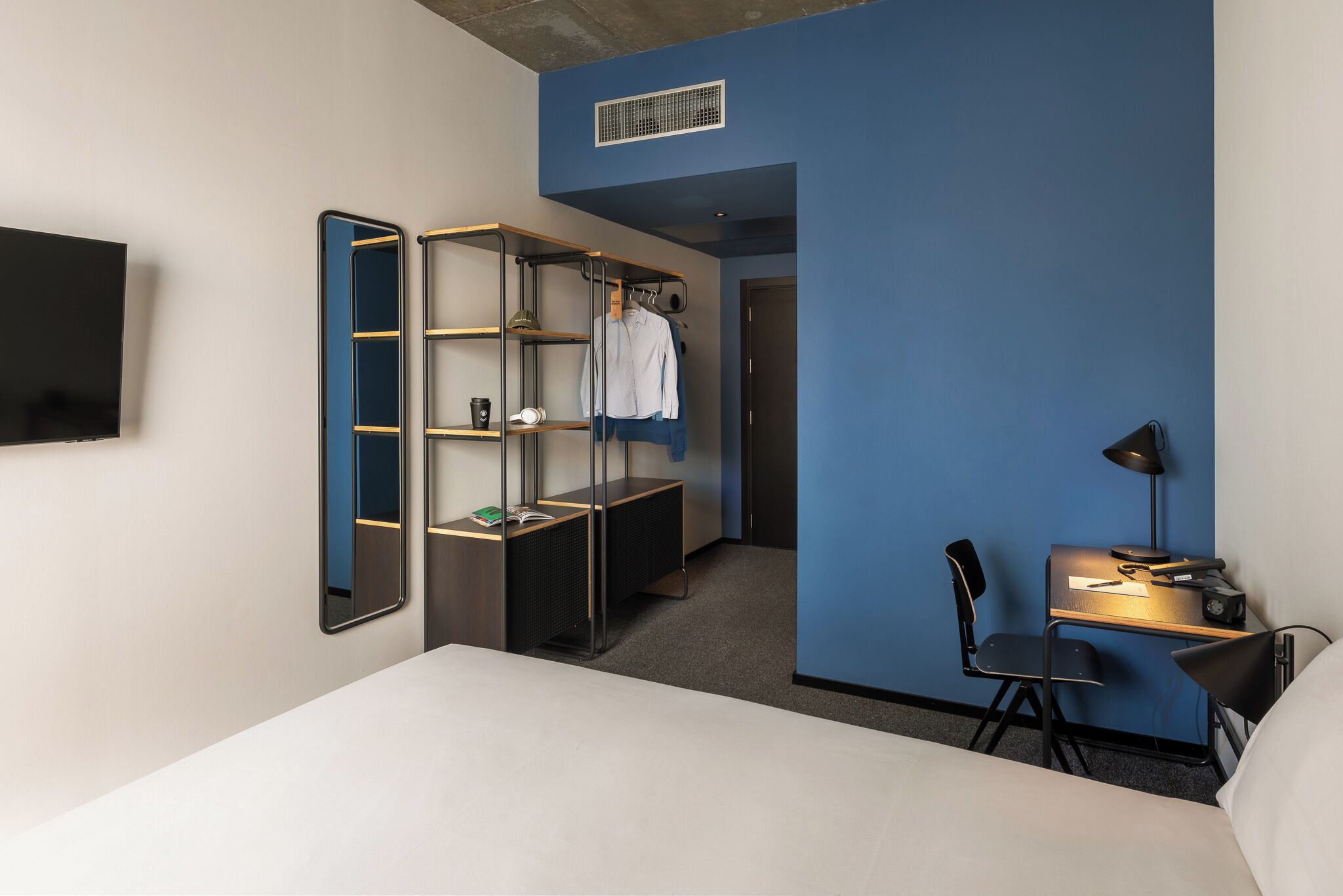 A hotel room with a bed, a desk with a chair, a wardrobe, a TV, and a shelf with a mirror.