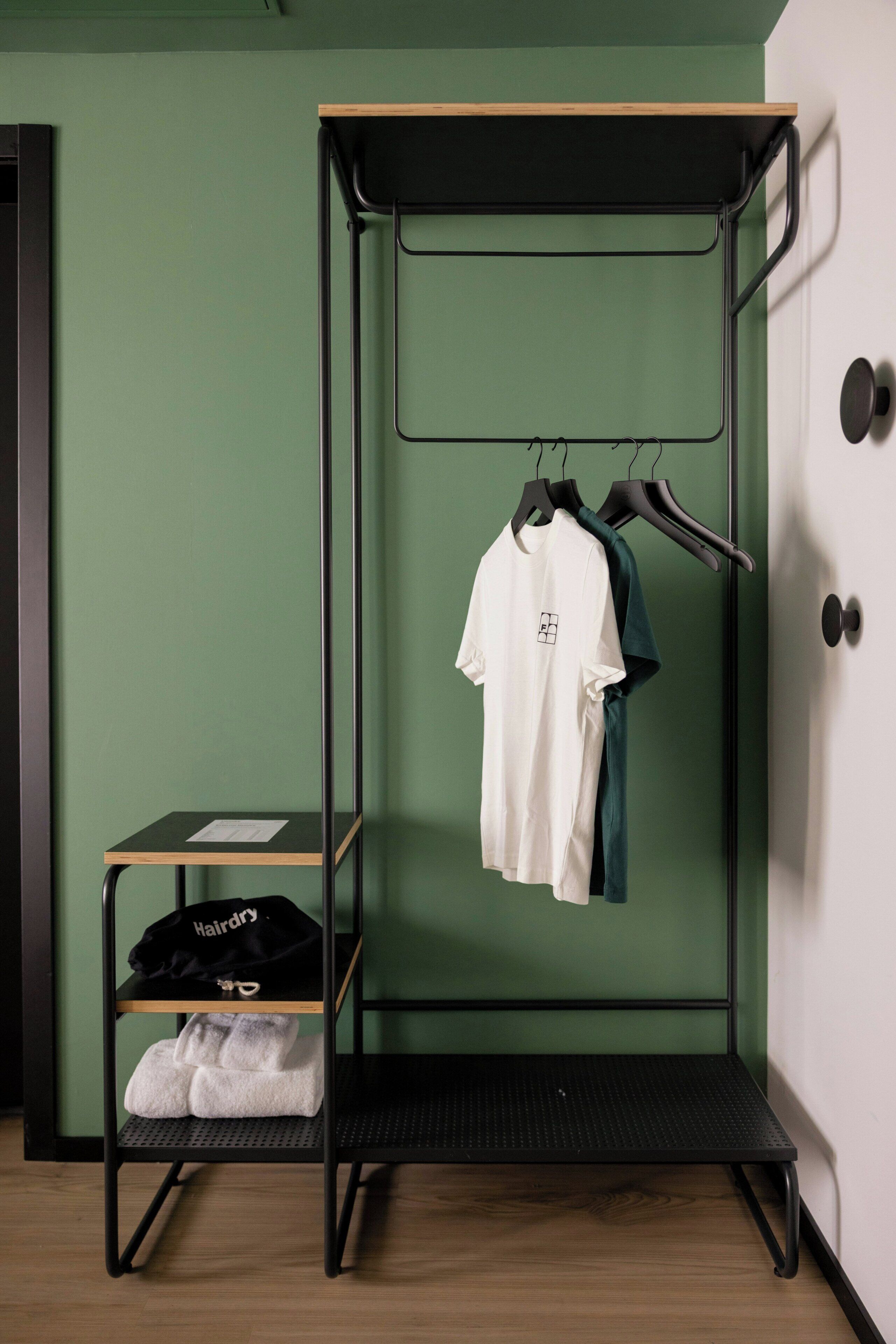 Una habitación compacta con una pared verde, un armario metálico negro con repisas, y una pequeña mesa con una camisa blanca y una gorra negra encima.