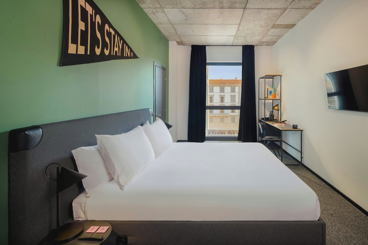 A hotel room with a large bed, a desk with a chair, a TV on the wall, and a window with a view of buildings.