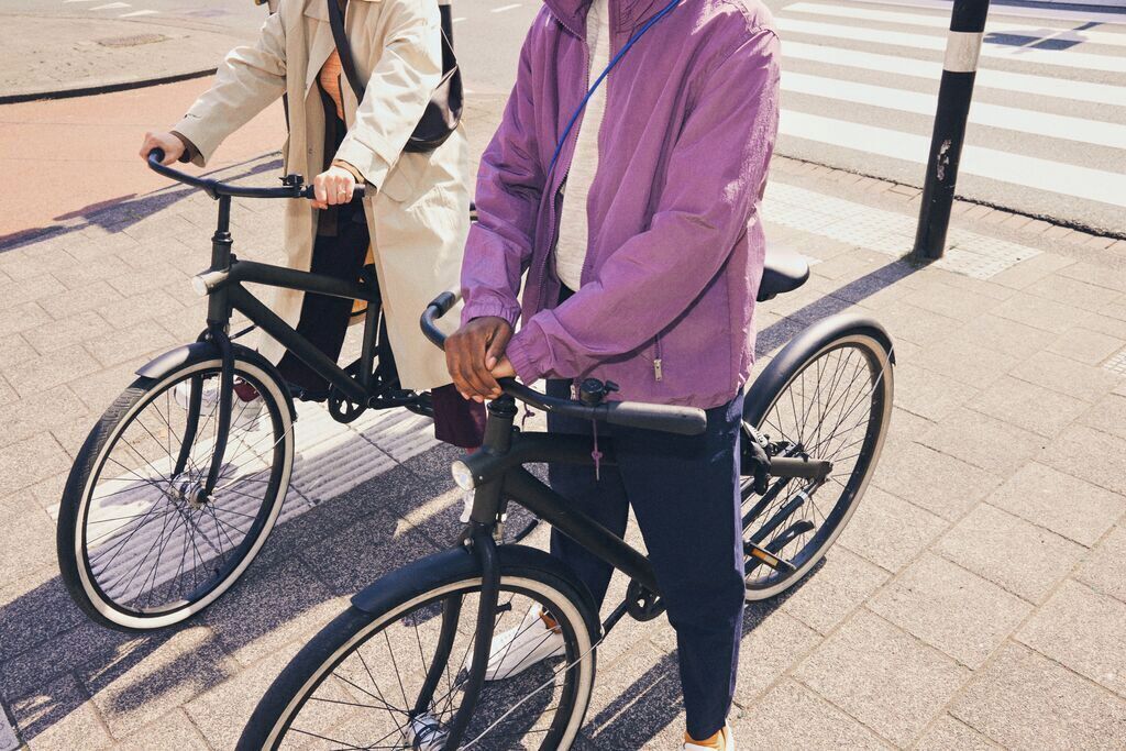 Deux personnes à vélo, l’une portant une veste violette et l’autre un manteau beige, se tiennent sur une surface pavée, avec un passage pour piétons en arrière-plan.