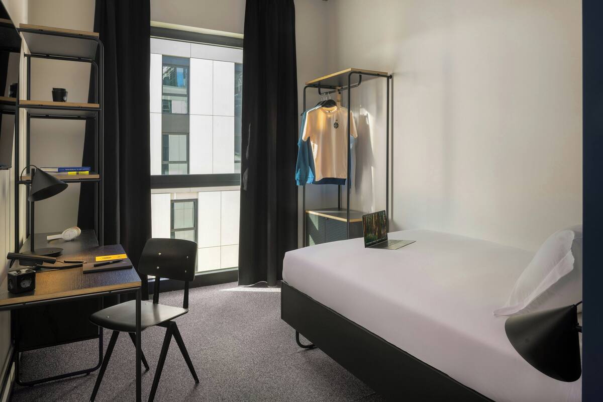 A hotel room with a bed, a desk with a chair, a bookshelf, and a window with curtains.
