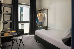 A hotel room with a bed, a desk with a chair, a bookshelf, and a window with curtains.
