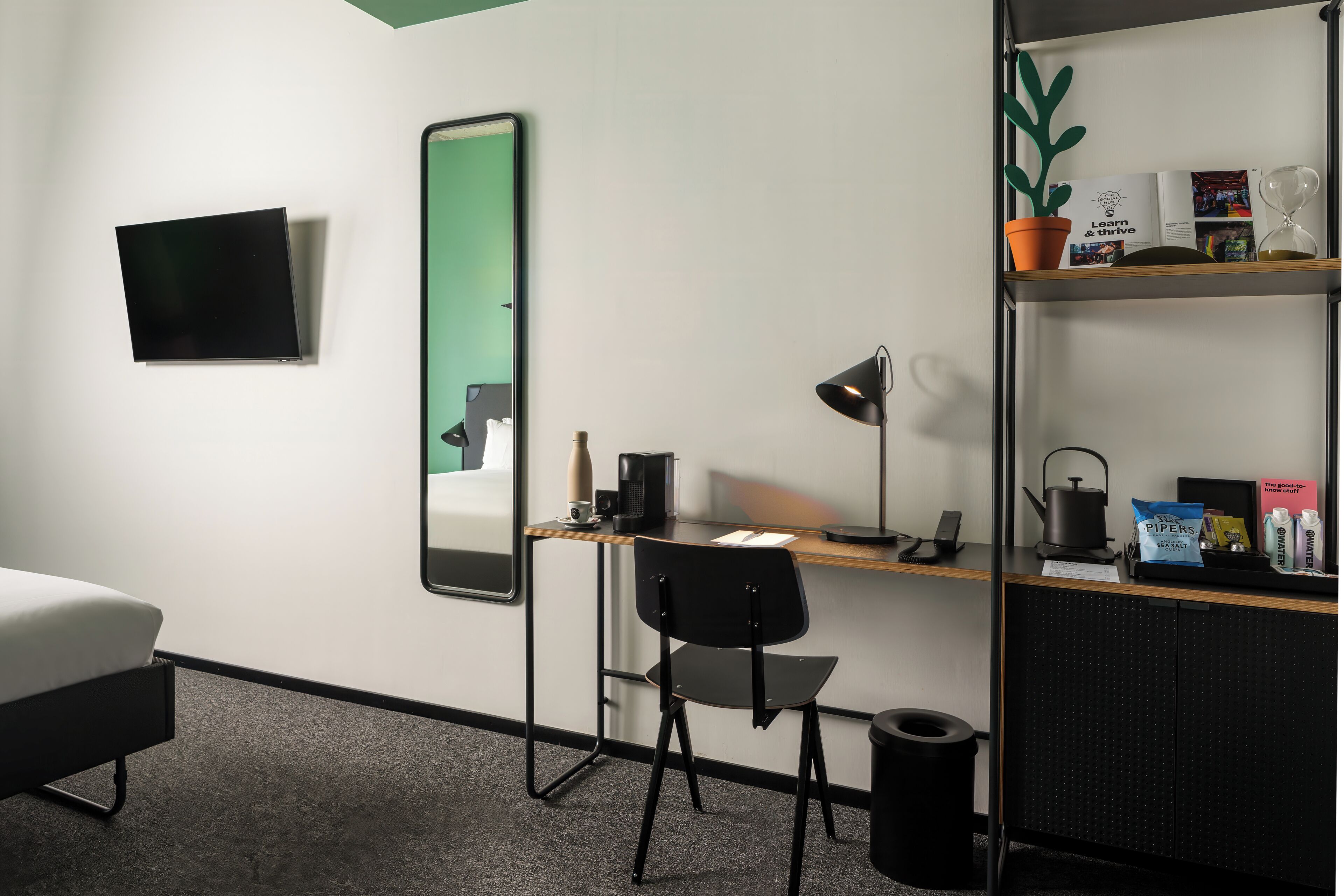 A hotel room with a bed, a desk with a chair, a mirror, a television, a shelf with books and a potted plant.