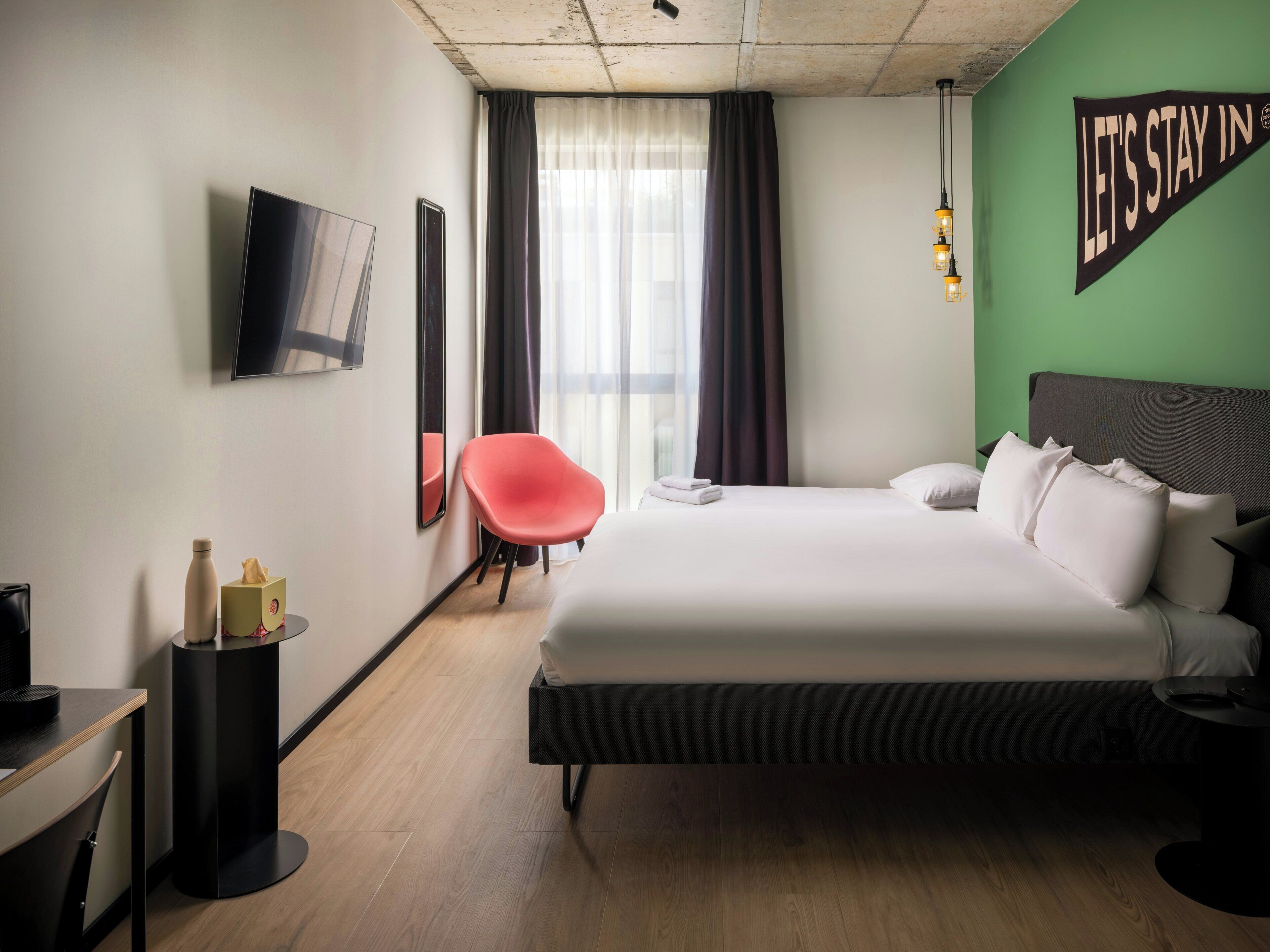 Ein Hotelzimmer mit Bett, Sessel, Fernseher und einem Schild mit der Aufschrift 'LET'S STAY IN'.