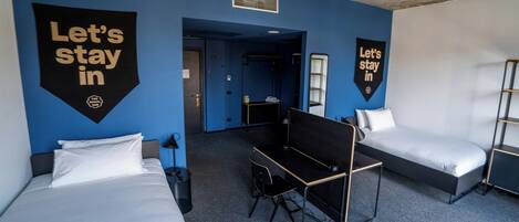 A hotel room with two beds, a desk, and a chair. There are two 'Let's stay in' banners on the walls.