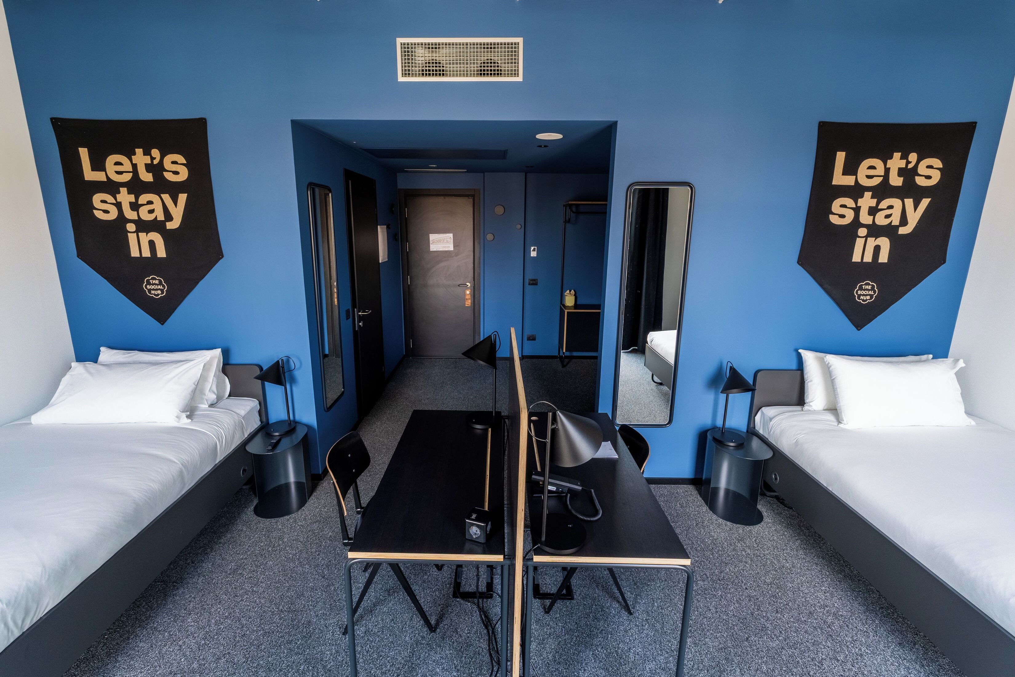 Quarto de hotel com duas camas, uma escrivaninha e uma cadeira. Há dois cartazes nas paredes com a inscrição 'Fique conosco'.