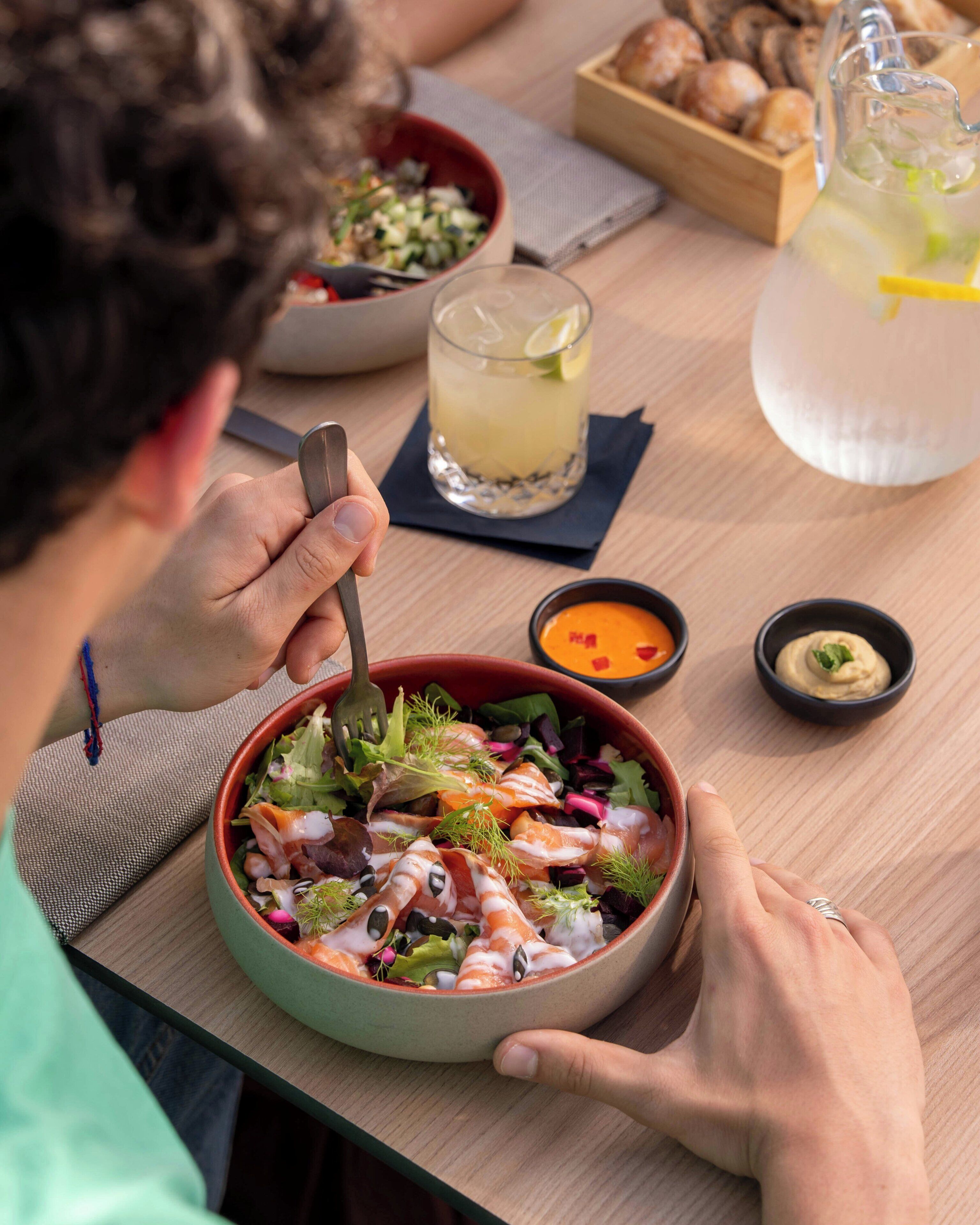 A person dining at a table with a bowl of salad, a drink, and small bowls of condiments.