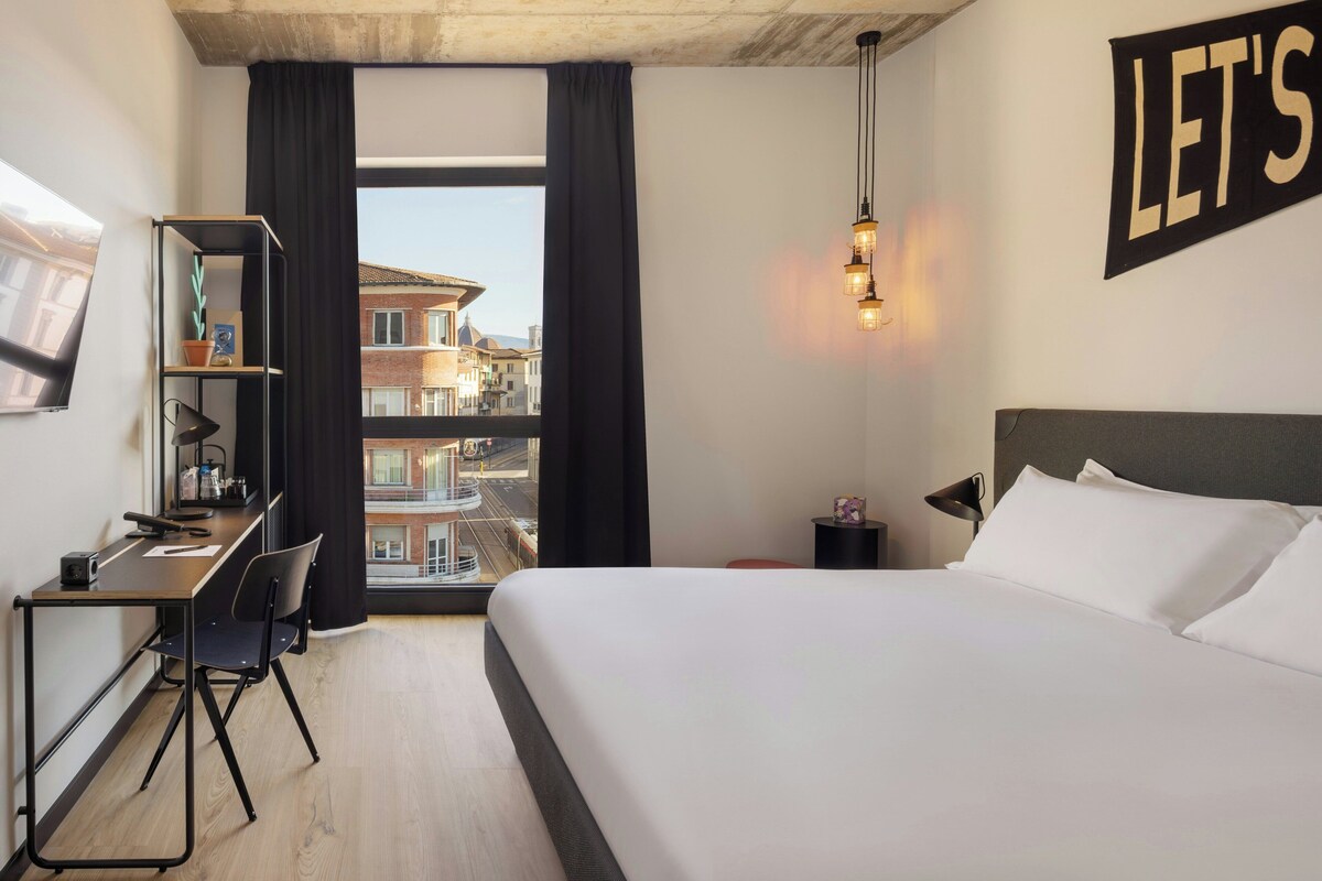 Habitación de hotel con cama, escritorio, silla y vista a edificios a través de una ventana.