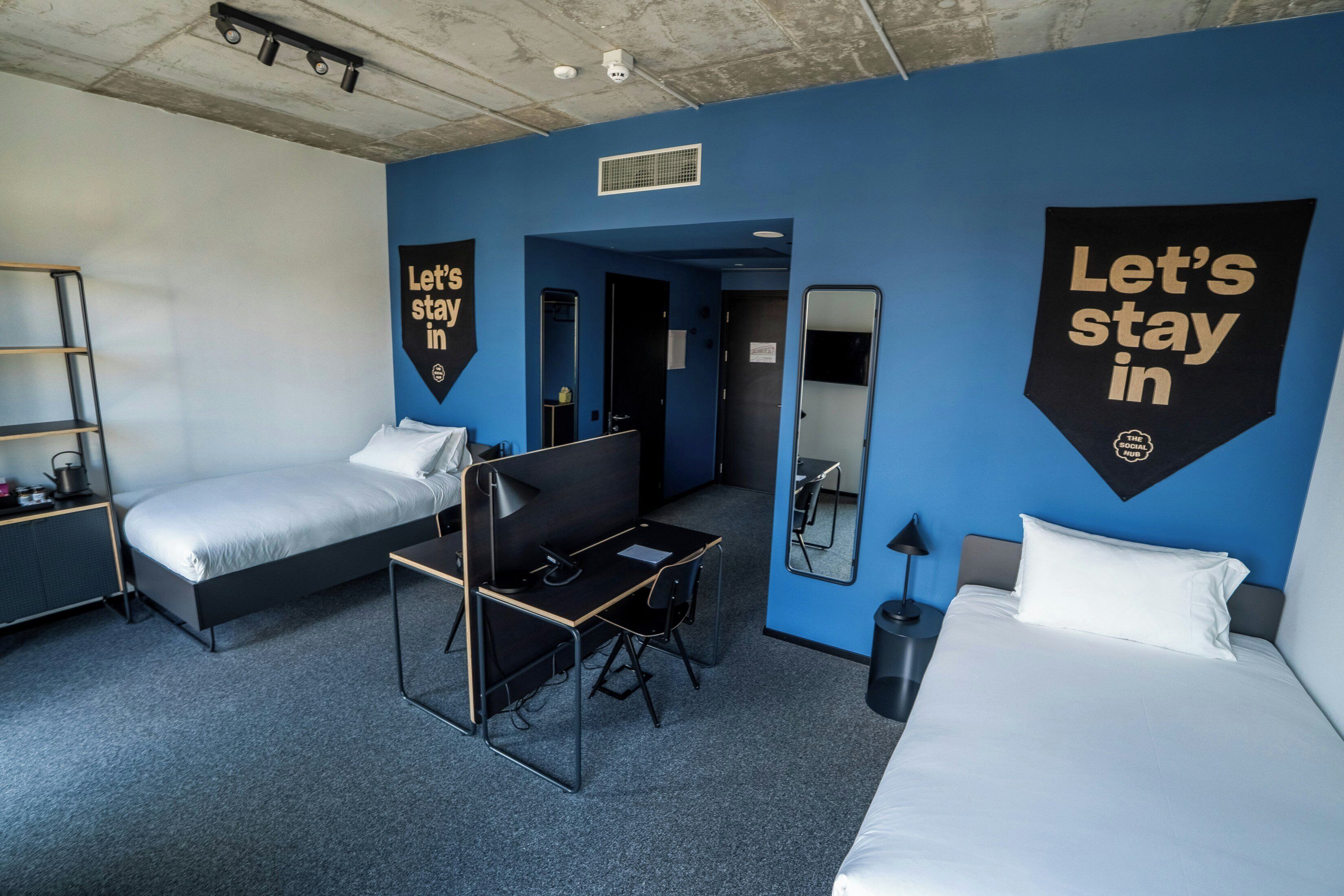 A hotel room with a bed, desk, chair, and a mirror. There are two large banners on the walls.