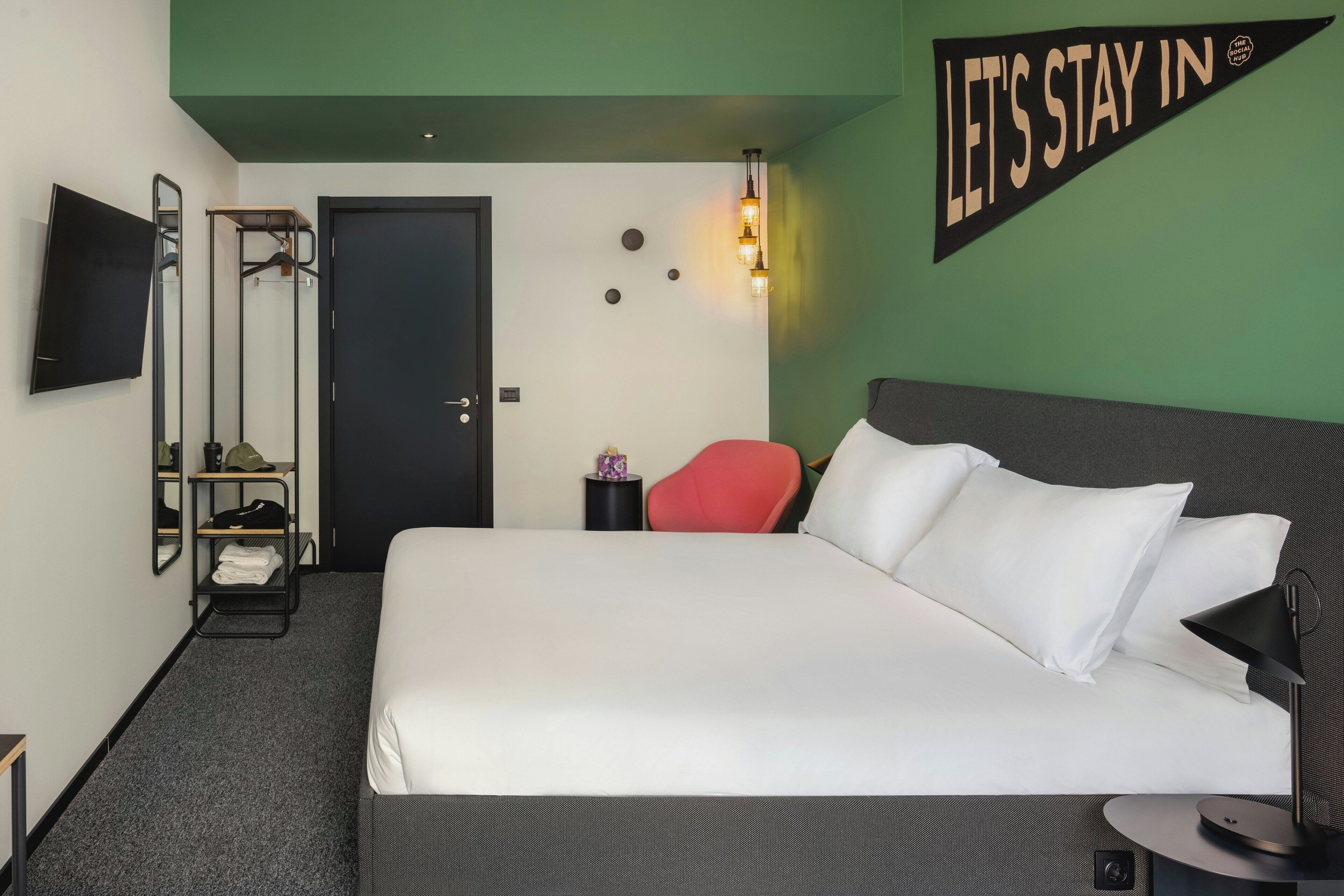 A hotel room with a bed, a TV, a red chair, a wall-mounted light, and a 'LET'S STAY IN' sign.