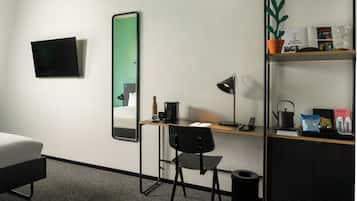 Habitación de hotel con cama, escritorio con silla, espejo, televisor, estante pequeño con una planta y un cesto de basura.