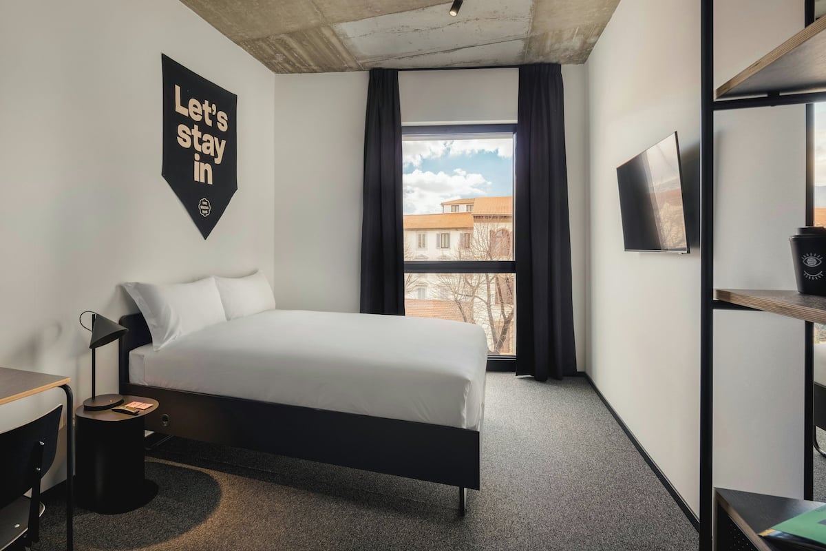 A modern hotel room with a bed, a desk, a chair, a TV, and a window with a view of buildings.