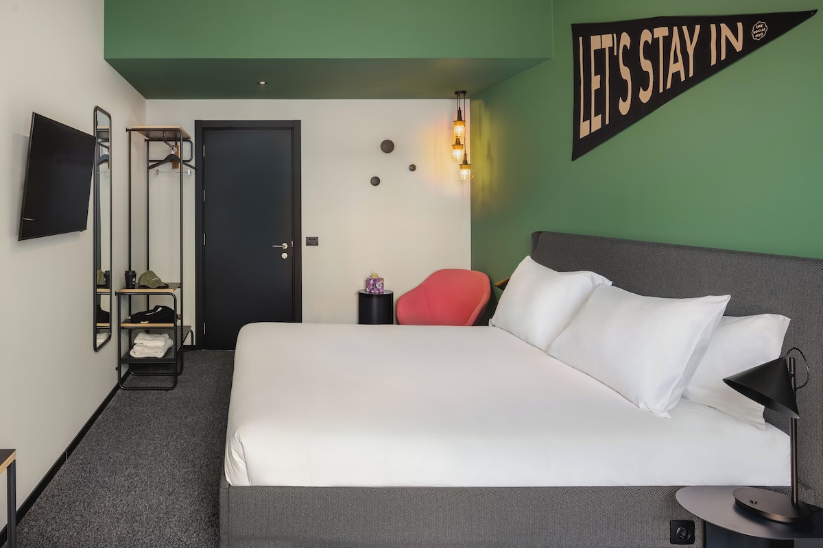A hotel room with a bed, a TV, a red chair, a coat rack, and a 'Let's Stay In' sign.