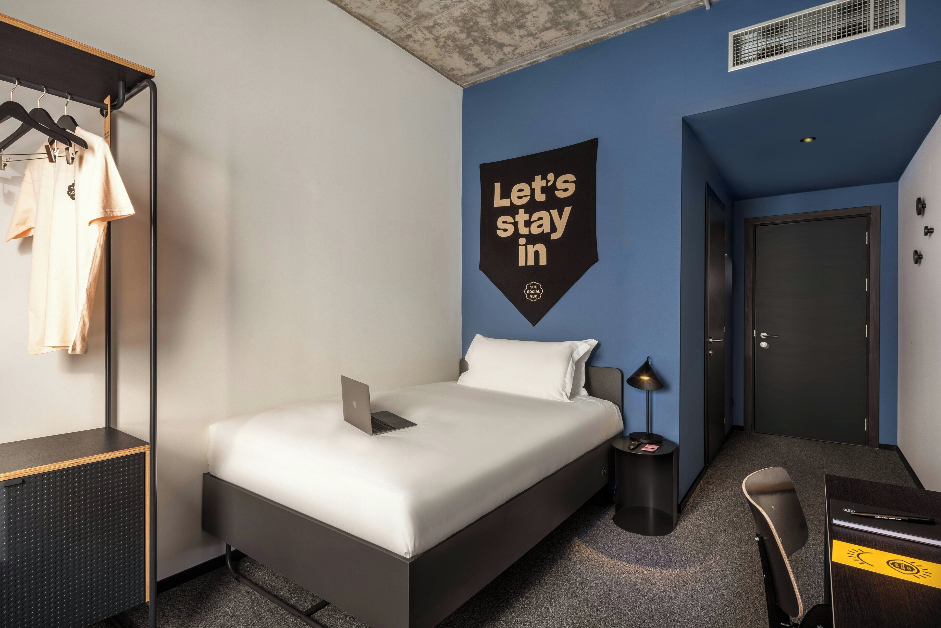 Ein modernes Hotelzimmer mit Bett, Schreibtisch, Stuhl und Kleiderschrank, sowie einem Wandzeichen.