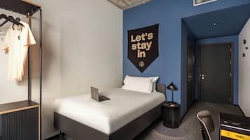 Ein modernes Hotelzimmer mit Bett, Schreibtisch, Stuhl und Kleiderschrank, sowie einem Wandzeichen.