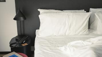 A neatly made bed with white bedding, a black lamp, a telephone, and a book on a red ottoman.