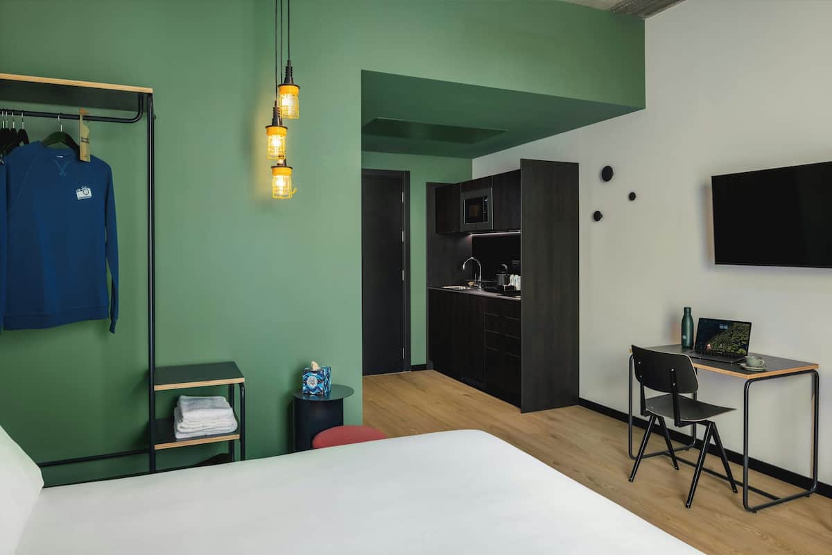A modern hotel room with a bed, a desk with a laptop, a kitchenette, and a wardrobe with clothes.