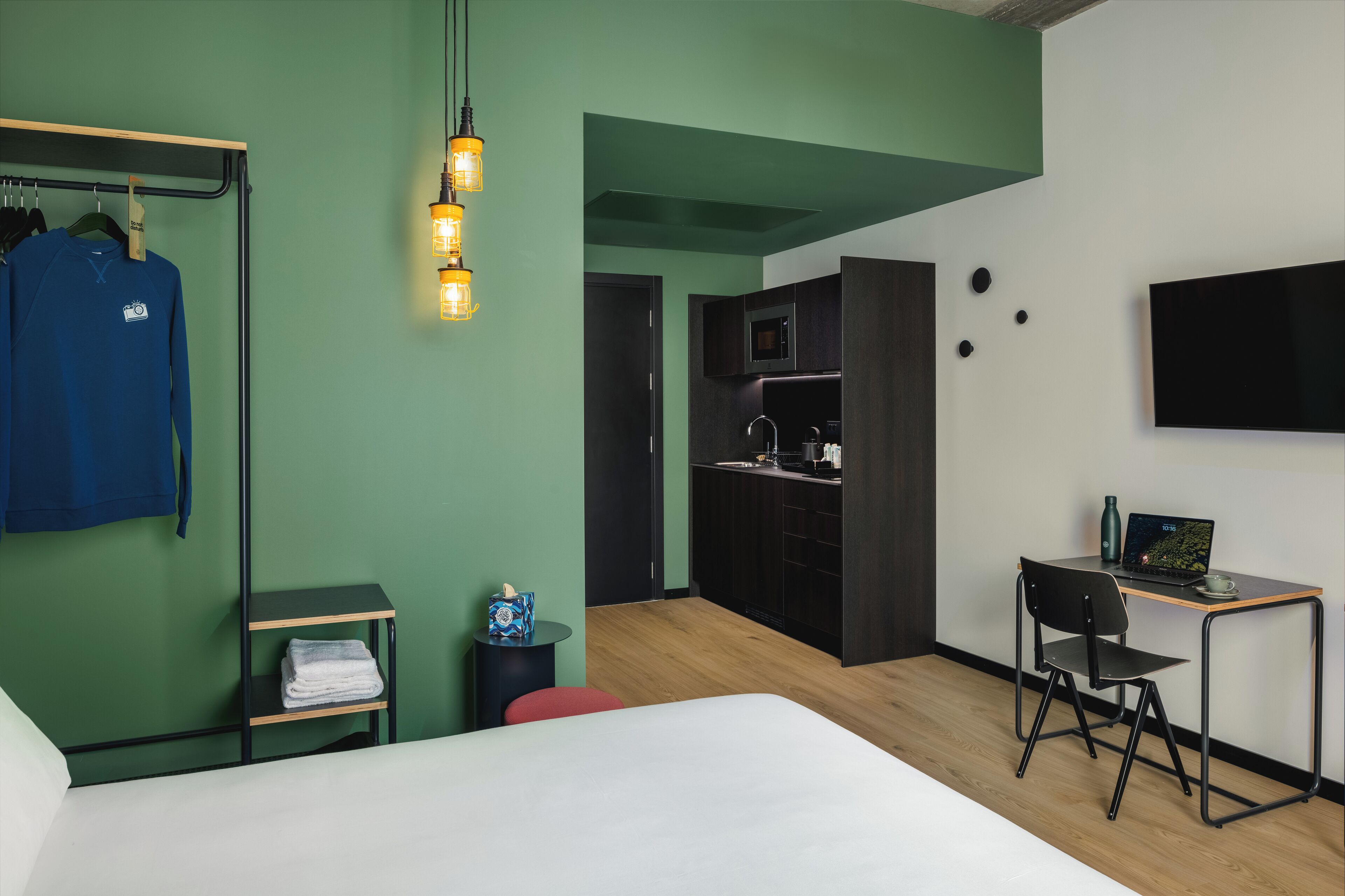 A modern hotel room with a bed, a desk with a laptop, a kitchenette, and a wardrobe with clothes hanging.