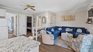 A living room with a blue sectional sofa, a white coffee table, and a patterned armchair. There is a ceiling fan, a TV mounted on the wall, and a door leading to another room. - Tower Shores Tiny Gem (Bethany Beach)