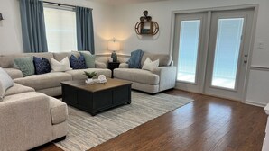 Un salon avec deux canapés beige, une table basse en bois foncé et un tapis. Il y a deux fenêtres équipées de stores, une porte et une étagère murale ornée d’objets décoratifs.