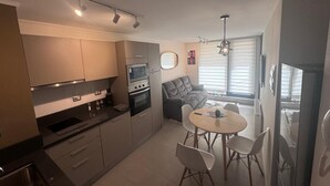 A compact kitchen with modern appliances, a dining area with a round table and chairs, and a grey sofa in a bright living space with large windows. - Painestone apartment (Puerto Natales)