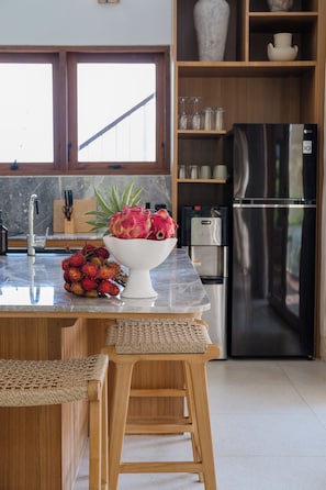 Una cocina con encimera de mármol, un bol de fruta, un refrigerador de acero inoxidable, estantes de madera y una ventana con vistas.