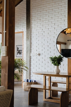 A room with a white brick wall, a round mirror, a wooden table, a bench, and a wicker basket with a plant.