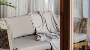A wicker sofa with a light-colored throw blanket, a wooden chair, and a potted plant in a room with sheer curtains. - Indah Sun Villa | Private Pool | 3BR | Pererenan, 2 Min to Cafés (Mengwi, Kabupaten Badung)