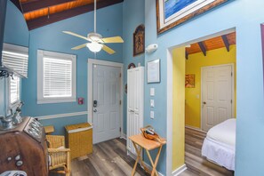 Una habitación con ventilador de techo, una puerta con detector de incendios, una silla de mimbre, una mesita pequeña y una cama con ropa blanca.