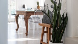 Ein modernes Interieur mit einem Holztisch, klaren Stühlen, einer Topfpflanze und einem gerahmten Kunstwerk auf einem Ständer.