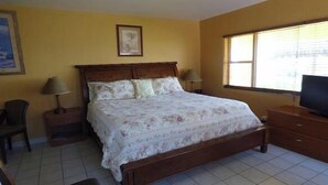 Une chambre à coucher avec un lit en bois, deux tables de chevet avec des lampes, un téléviseur posé sur une commode en bois et une fenêtre avec des stores.
