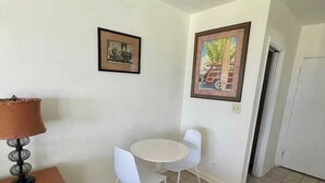 A small, clean room with a round table and two white chairs, a lamp, and two framed pictures on the walls. - King Studio with Sofa Sleeper, Full Kitchen,Ocean Views (Pompano Beach)