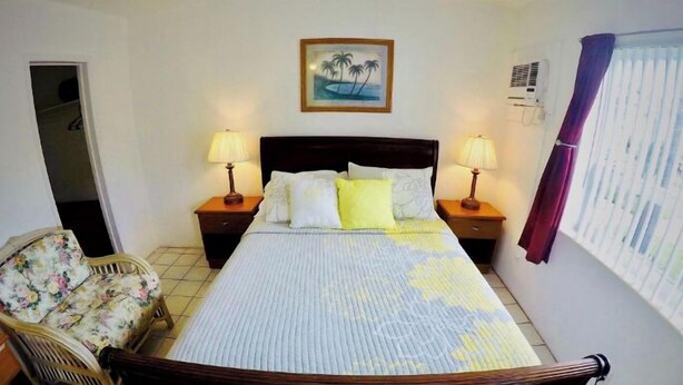 A hotel room with a bed, two bedside tables with lamps, a chair, a painting on the wall, and an air conditioning unit. - Cozy Queen Studio with Kitchen and Scenic Beach Views (Pompano Beach)