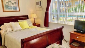 A hotel room with a wooden bed, a TV on a wooden side table, a lamp, and a window with curtains. - Cozy Queen Studio with Kitchen and Scenic Beach Views (Pompano Beach)