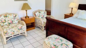 A bedroom with a wooden bed, two floral armchairs, a small bedside table with a lamp, and a nightstand with a lamp. - Cozy Queen Studio with Kitchen and Scenic Beach Views (Pompano Beach)