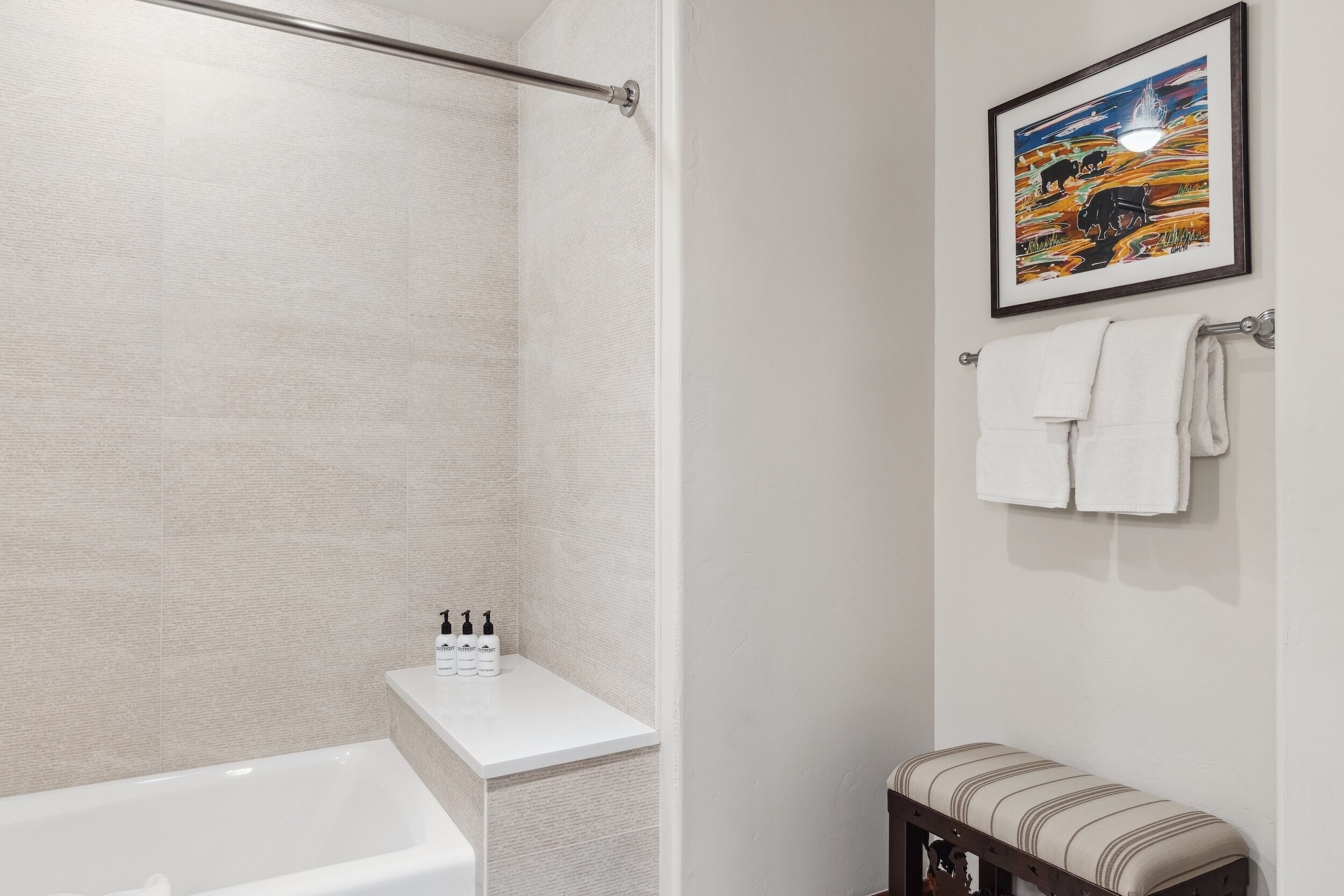 Een badkamer met een bad, douche en een bankje. Handdoeken hangen aan een rek en er is een lijst met een foto aan de muur.