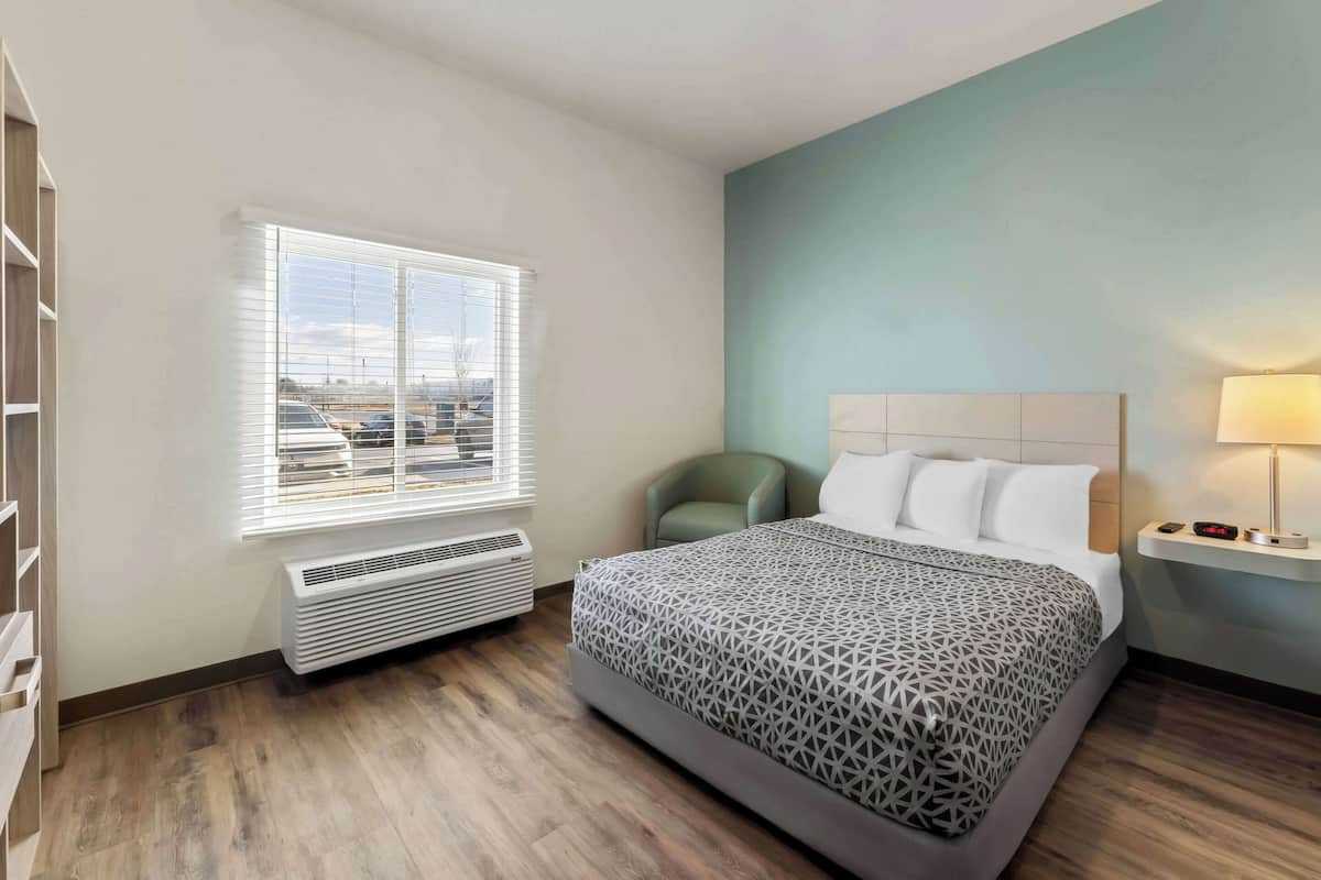 Une chambre d’hôtel avec un lit, une fenêtre avec des stores, un radiateur, un fauteuil vert et une table de chevet avec une lampe.