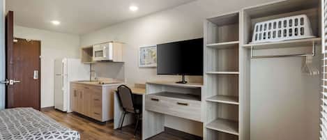 Habitación pequeña con cama, escritorio con computadora, microondas, refrigerador y armario con repisas.
