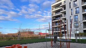 Leikkikenttä, jossa on hiekkaa ja oransseja sylinterimäisiä elementtejä, ja jonka ympärillä on nurmikkoalue sekä useita kerroksia sisältävä rakennus, jossa on parvekkeita.