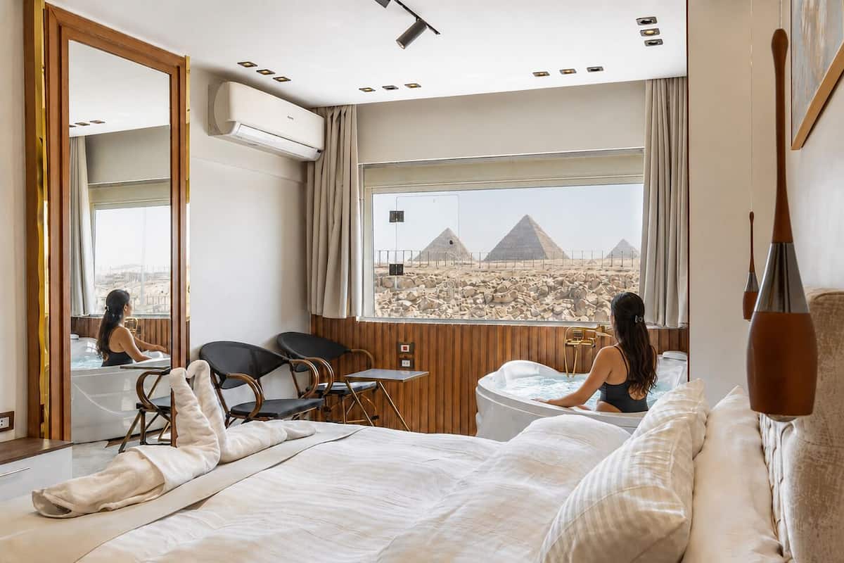 A hotel room with a large bed, a sitting area with chairs, a view of pyramids, and an air conditioning unit.