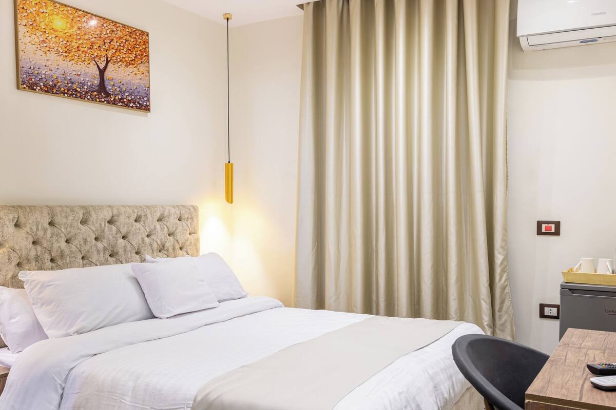 A hotel room with a bed, a desk, a chair, a painting, a hanging light, and a curtain.