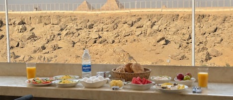 A breakfast table with various food items and a bottle of water, overlooking a desert landscape with pyramids.