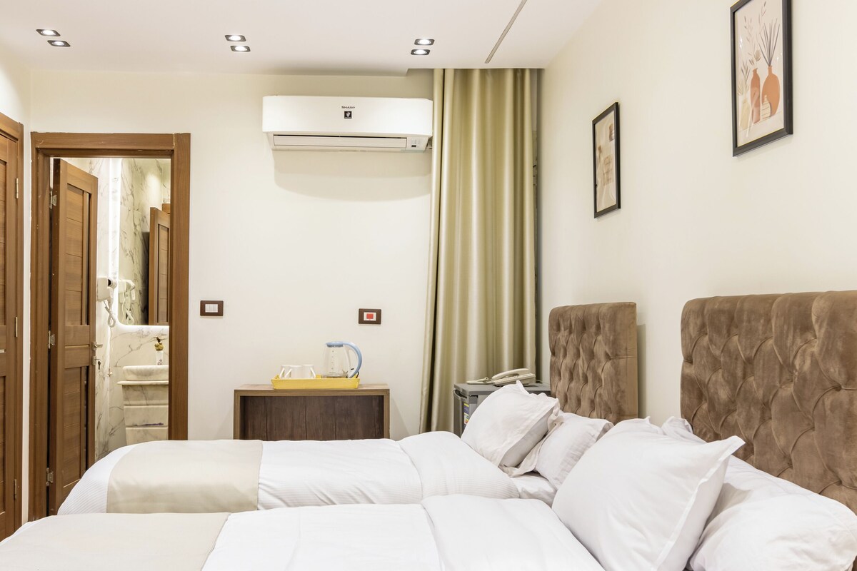 A hotel room with a bed, a bedside table with a phone, a mirror, an air conditioning unit, and a window with curtains.