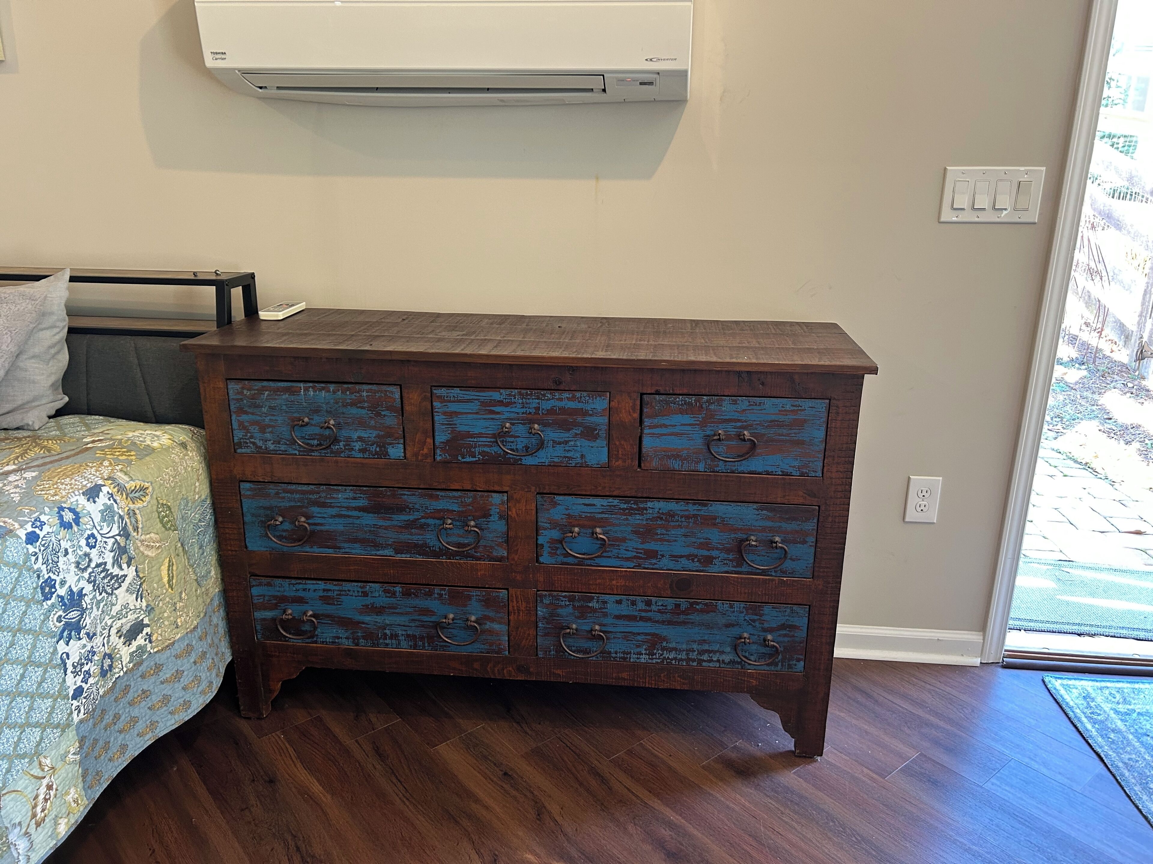 A wooden chest of drawers with blue paint, a bed with floral bedding, and a wall-mounted air conditioner.