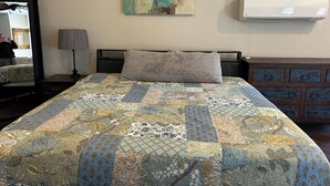A bed with a quilt, a bedside lamp, a mirror, a wall-mounted air conditioner, and a dresser with drawers.