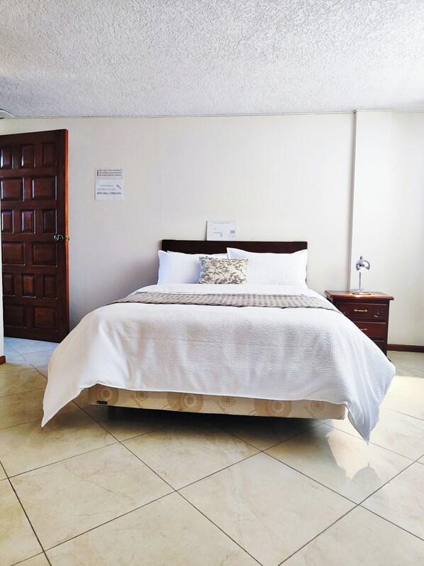 A neatly made bed with a white comforter and a patterned pillow, a wooden nightstand with a lamp, and a door with a sign indicating hotel administration contact. - Hotel Sweet Garden (Cumbayá)